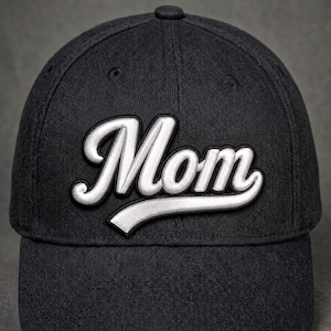 May include: A dark charcoal gray baseball cap featuring the word Mom embroidered in white script lettering across the front panels. The embroidery has a raised, three-dimensional appearance with a black outline, and the cap has a structured crown.