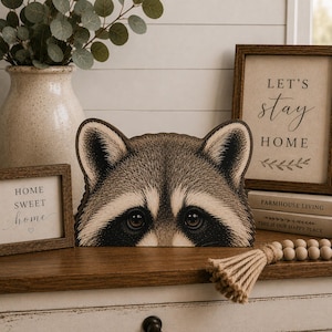 May include: A flat, wooden cutout shaped like the head of a raccoon, featuring detailed brown, black, and cream-colored fur patterns, pointed ears, and dark, expressive eyes, designed as a decorative tabletop piece for home display.