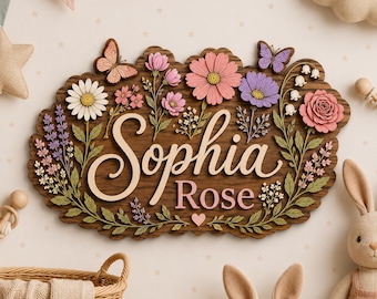 以下が含まれることがあります： 木製のウォールサインで、Sophia Roseという名前が浮き彫りになっています。ピンク、紫、白の色合いのデイジー、バラ、蝶などの重なり合った花の切り抜きと、緑の葉のアクセントで装飾されています。
