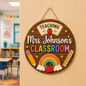 Può includere: Un cartello circolare in legno per aula con finitura a venature scure, che riporta il testo "TEACHING" e "Mrs. Johnson's CLASSROOM". Include una matita gialla, un righello in legno, un arcobaleno colorato e icone di mele rosse con stelle gialle.
