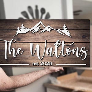 Könnte beinhalten: Holzschild mit einem Berg- und Baumdesign. Das Schild zeigt den Text "The Wattons est 2025" in weißer Schrift. Der Hintergrund ist eine dunkelbraune Holzmaserung.