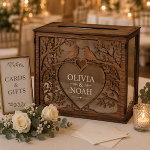 May include: A rectangular dark brown wooden card box with a slot on top, featuring a laser-cut tree and bird design surrounding a central heart-shaped window. The glass is etched with the names Olivia & Noah in white lettering.