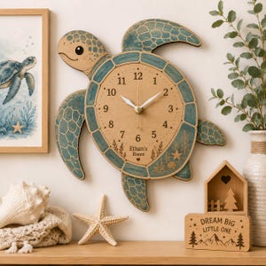 May include: A wall clock shaped like a sea turtle with a light wood face, blue-toned shell edges, and white clock hands. The clock face features black numbers from 1 to 12, the text Ethan's Room, and small illustrations of seaweed, a starfish, and a shell.