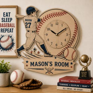 Könnte beinhalten: Eine hölzerne Wanduhr in Form eines Baseballs mit einer Spielerfigur, die ein Trikot mit der Aufschrift MASON 27 trägt. Das Zifferblatt hat schwarze Zahlen und Zeiger. Darunter zeigt ein Banner den Text MASON'S ROOM mit gekreuzten Schlägern.