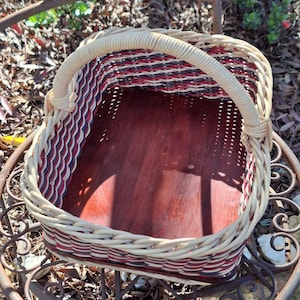 May include: A rectangular basket featuring a smooth, dark reddish-brown wooden base and woven sides. The sides are constructed with a pattern of thin, alternating strips in black, white, and red, finished with a thick, light beige woven rim and handle.