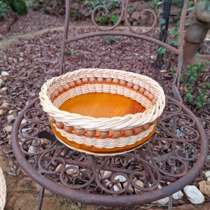 May include: A round basket featuring a smooth, polished wooden base and woven light-coloured wicker sides. The design includes a decorative row of small, round wooden beads integrated into the weave along the upper rim of the basket structure.