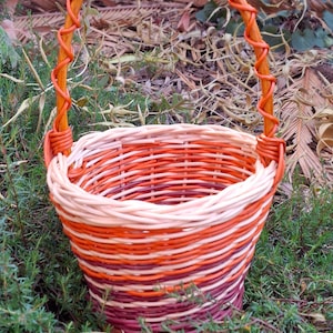 May include: A hand-woven basket with a handle, displaying a colour gradient from light beige to deep burgundy. The basket's body is intricately woven, and the handle is wrapped in orange-toned material. The basket is set against a backdrop of green foliage.