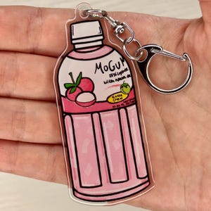 May include: A clear acrylic keychain in the shape of a pink lychee drink bottle. The keychain has a silver clasp and a ring. The bottle design includes the text "Mogu" and "Gotta Chew", along with lychee fruit illustrations.