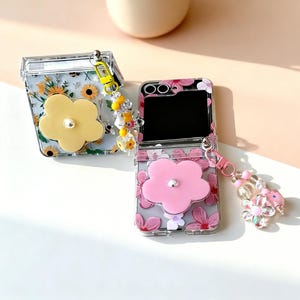 Puede incluir: Dos fundas transparentes para teléfono con diseños florales. Una funda presenta una flor amarilla y un patrón de girasoles, la otra un diseño de flores rosas. Ambas fundas tienen colgantes de cuentas decorativas.