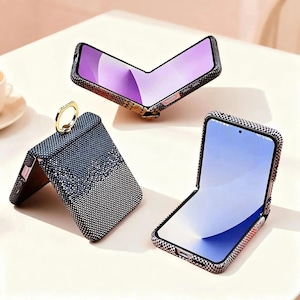 May include: Three open foldable smartphones with various patterned cases. One case is black with a gold ring, another has a black and white chequered pattern, and the third has a snakeskin pattern. The phone screens show a light blue and purple gradient.