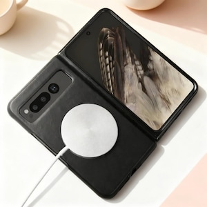 May include: Black foldable smartphone with a silver wireless charger attached. The phone is open, displaying a screen with a feather design. The charger is round and connected by a white cable.
