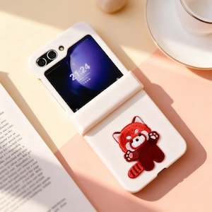 May include: A white phone case with a red panda patch. The phone case is open, revealing a phone screen displaying the time 21:24. The case is on a light pink surface.