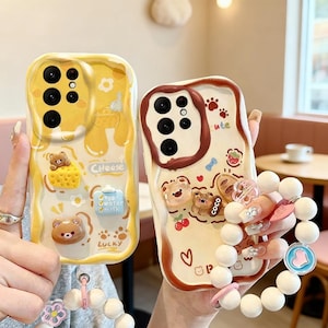 Puede incluir: Dos fundas de teléfono con diseños de osos lindos. Una es amarilla con gráficos de queso y huevo, la otra es de color crema con huellas de patas y una fresa. También se ve una pulsera de cuentas blancas con detalles en rosa.