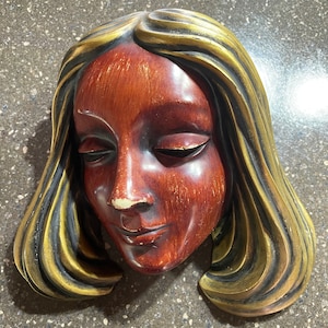 May include: A wooden wall sculpture of a woman's face with long, flowing hair. The hair is painted gold and black, and the face is painted a reddish brown.