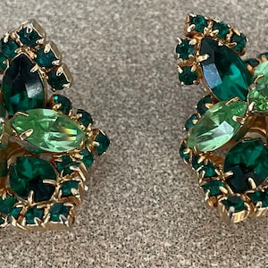Pode incluir: Um par de brincos vintage de encaixe com um design floral. Os brincos são adornados com strass verde esmeralda e verde claro em metal dourado. Os brincos têm uma forma quadrada com um aglomerado de pedras.