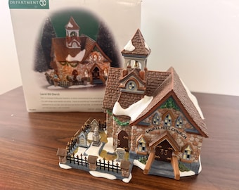 May include: A miniature ceramic Laurel Hill Church building with a stone-textured facade, brown shingled roof with white snow accents, a bell tower, and a small graveyard enclosed by a black metal fence, accompanied by its original cardboard box.