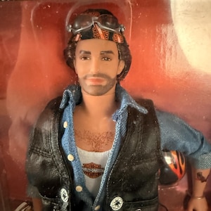 May include: A male doll with brown hair and a beard, wearing a black bandana with orange text, goggles, a blue denim shirt, a white tank top with a Harley-Davidson logo, and a black vest with silver buttons, packaged in a clear plastic box.