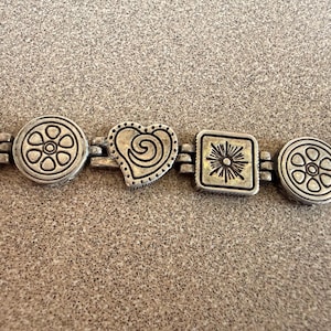 Puede incluir: Pulsera plateada con formas alternas: cuadrados con dise&ntilde;os de sol, patrones florales circulares y formas de coraz&oacute;n con detalles de espiral. La pulsera est&aacute; unida por peque&ntilde;os eslabones rectangulares.