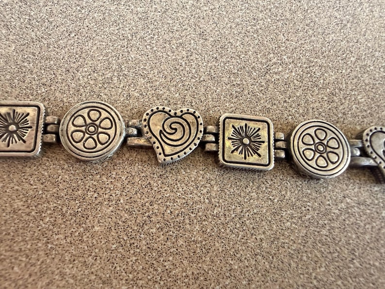 Puede incluir: Pulsera plateada con formas alternas: cuadrados con dise&ntilde;os de sol, patrones florales circulares y formas de coraz&oacute;n con detalles de espiral. La pulsera est&aacute; unida por peque&ntilde;os eslabones rectangulares.