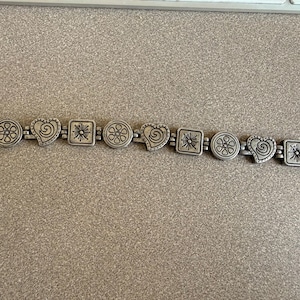 Puede incluir: Pulsera plateada con formas alternas: c&iacute;rculos con dise&ntilde;os florales, corazones con espirales y cuadrados con dise&ntilde;os de sol. La pulsera tiene un cierre de palanca y un peque&ntilde;o dije de coraz&oacute;n.