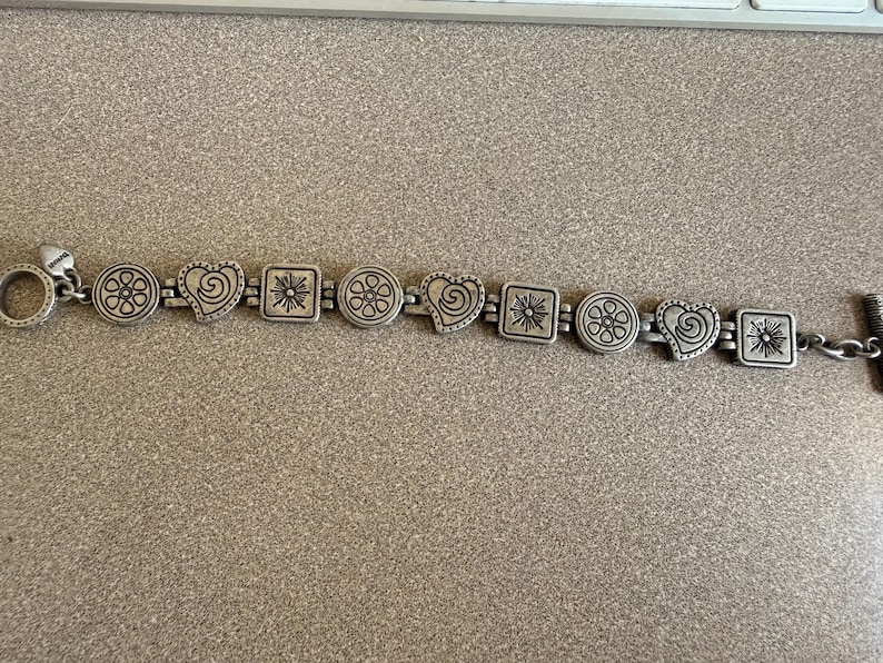 Puede incluir: Pulsera plateada con formas alternas: c&iacute;rculos con dise&ntilde;os florales, corazones con espirales y cuadrados con dise&ntilde;os de sol. La pulsera tiene un cierre de palanca y un peque&ntilde;o dije de coraz&oacute;n.