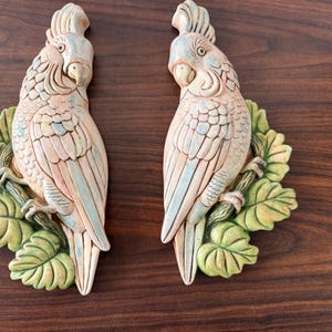 May include: Two matching wall decorations featuring detailed parrot sculptures. The parrots are cream-colored with blue and pink accents, perched on green leafy branches. The sculptures are made of a solid material and are mounted on a dark wood surface.