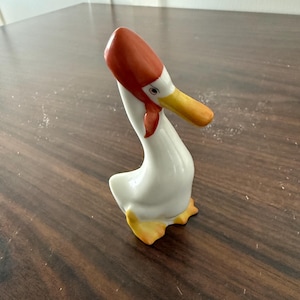 May include: A ceramic goose figurine with a white body, orange hat, and yellow beak and feet. The goose has a red scarf around its neck and is posed in a standing position. The figurine is on a dark brown wooden surface.