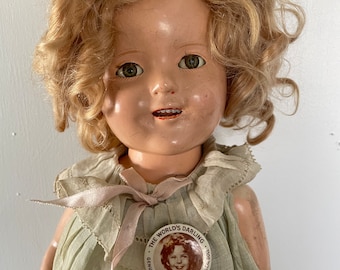 May include: A vintage Shirley Temple doll with blonde curly hair, wearing a light green dress with a white lace collar and a pink ribbon. The doll has a button on her dress with a picture of Shirley Temple and the text "The World's Darling - Genuine Shirley Temple -  The Ideal Doll."