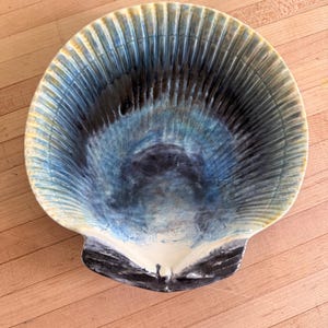 May include: A ceramic bowl shaped like a large scallop shell, featuring a gradient glaze that transitions from a deep navy blue center to lighter shades of blue and cream along the ribbed, scalloped edges, with a smooth, glossy finish throughout.