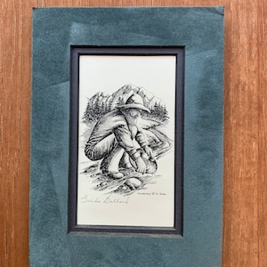 Puede incluir: Un dibujo en blanco y negro de una persona buscando oro en un arroyo. El dibujo está enmarcado en un paspartú azul oscuro y un marco negro. La firma "Zelda Ballard" es visible en la esquina inferior izquierda del dibujo.