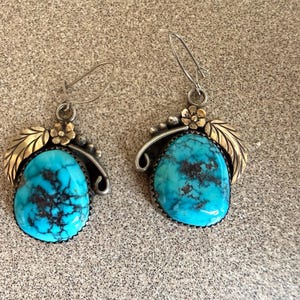 Pode incluir: Um par de brincos pendentes com grandes pedras de turquesa em forma de gota com veios escuros. Cada brinco é decorado com uma pequena flor de metal dourado e um design de folha curva dourada, suspensos por um gancho prateado simples.