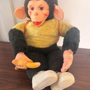 Può includere: Una bambola scimmia di peluche con viso, orecchie e mani in plastica stampata. Indossa una maglietta gialla con bretelle rosse e pantaloni neri. La scimmia ha pelliccia nera sulla testa e sulle braccia e tiene una banana di plastica in mano.