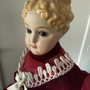 May include: A vintage porcelain doll with a serene expression. The doll has blonde, curly hair and is dressed in a burgundy velvet dress with a white lace collar and bow. The doll's face is delicately painted with detailed features.