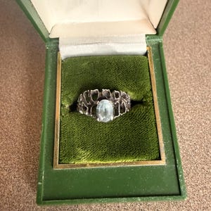 Puede incluir: Un anillo de plata con una piedra preciosa ovalada de color azul claro, engastada en un diseño calado texturizado. El anillo se exhibe en un joyero verde vintage con ribete dorado e interior de terciopelo verde.