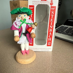 May include: A Steinbach wooden figurine of a traditional German nutcracker. The figurine has a green hat, pink coat, and white lace collar. It stands on a wooden base, with a box in the background.