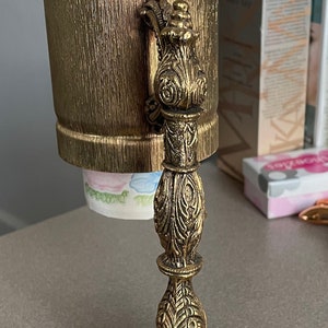 Puede incluir: Un soporte decorativo de metal dorado con una parte superior redonda y tres patas. El soporte presenta un diseño ornamentado y texturizado con motivos florales. La parte superior del soporte tiene un recipiente cilíndrico con un acabado dorado texturizado.