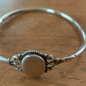 Op de afbeelding: Een zilveren armband met een decoratieve ovale hanger. De hanger heeft een verhoogd, versierd ontwerp rond de rand.