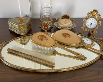 May include: A gold-toned vanity set with a mirrored tray, two hand mirrors, a hairbrush, a comb, a small jewelry box, a soap dish with a bar of soap, and a small clock. The set features decorative accents with amber-colored glass stones.