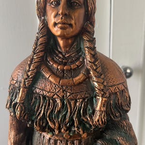 May include: A copper-colored statue of a Native American woman with long braided hair. She is wearing a beaded necklace and a fringed garment. The statue is detailed and realistic.