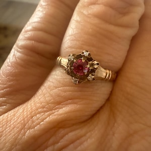 Op de afbeelding: Een gouden ring met een centrale rode edelsteen omringd door kleinere witte edelstenen. De ring heeft een gedetailleerd, sierlijk ontwerp met een vintage uitstraling. De ring zit om een vinger.