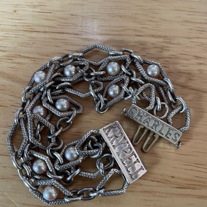 May include: A silver chain bracelet with multiple strands and pearl accents. The bracelet has two silver clasps with the words "CHARLES" and "KRUPEL" engraved on them.