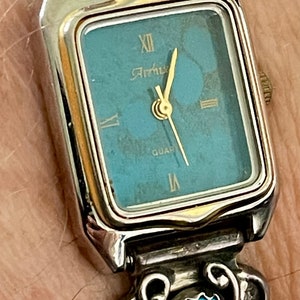 Op de afbeelding: Een zilveren horloge met een turquoise steen op de band en een vierkante wijzerplaat met een blauwe wijzerplaat. De wijzerplaat van het horloge zegt "Artnus" en "Quartz".