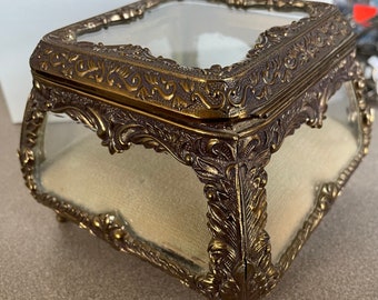 May include: A gold-toned glass trinket box with ornate detailing and a clear glass lid. The box has a velvet lining and sits on four small feet.