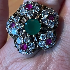 Puede incluir: Un anillo grande y adornado con una piedra verde rodeada de piedras más pequeñas transparentes y rosadas.