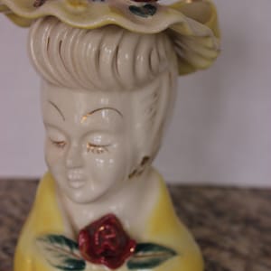 Könnte beinhalten: Eine antike Keramikfigur einer Frau mit einem gelben Hut und einer roten Rose. Die Figur sitzt und hat ein weißes Gesicht mit geschlossenen Augen.
