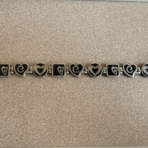 Puede incluir: Pulsera plateada con dijes alternos en forma de coraz&oacute;n y cuadrados con centros de esmalte negro. Los dijes est&aacute;n conectados por una cadena y tienen dise&ntilde;os de coraz&oacute;n. La pulsera tiene un cierre de palanca.