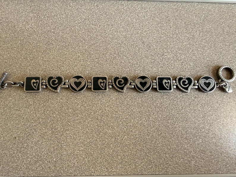 Puede incluir: Pulsera plateada con dijes alternos en forma de coraz&oacute;n y cuadrados con centros de esmalte negro. Los dijes est&aacute;n conectados por una cadena y tienen dise&ntilde;os de coraz&oacute;n. La pulsera tiene un cierre de palanca.