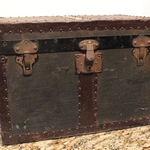 May include: A vintage, dark brown leather-covered trunk with metal accents and two brass clasps. The trunk has a black painted trim and is secured with a metal latch.