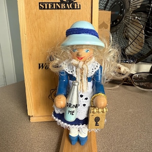 May include: A wooden Steinbach nutcracker doll of a character in a blue dress and hat, holding a bottle that says "DRINK ME" and a small purse. The doll has blonde hair and is standing on a wooden base.