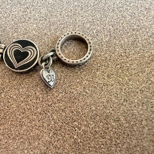 Puede incluir: Primer plano de una pulsera plateada con dijes en forma de coraz&oacute;n. La pulsera presenta un eslab&oacute;n circular con peque&ntilde;os detalles de puntos y un dije en forma de coraz&oacute;n con una inicial. La pulsera est&aacute; sobre una superficie texturizada.